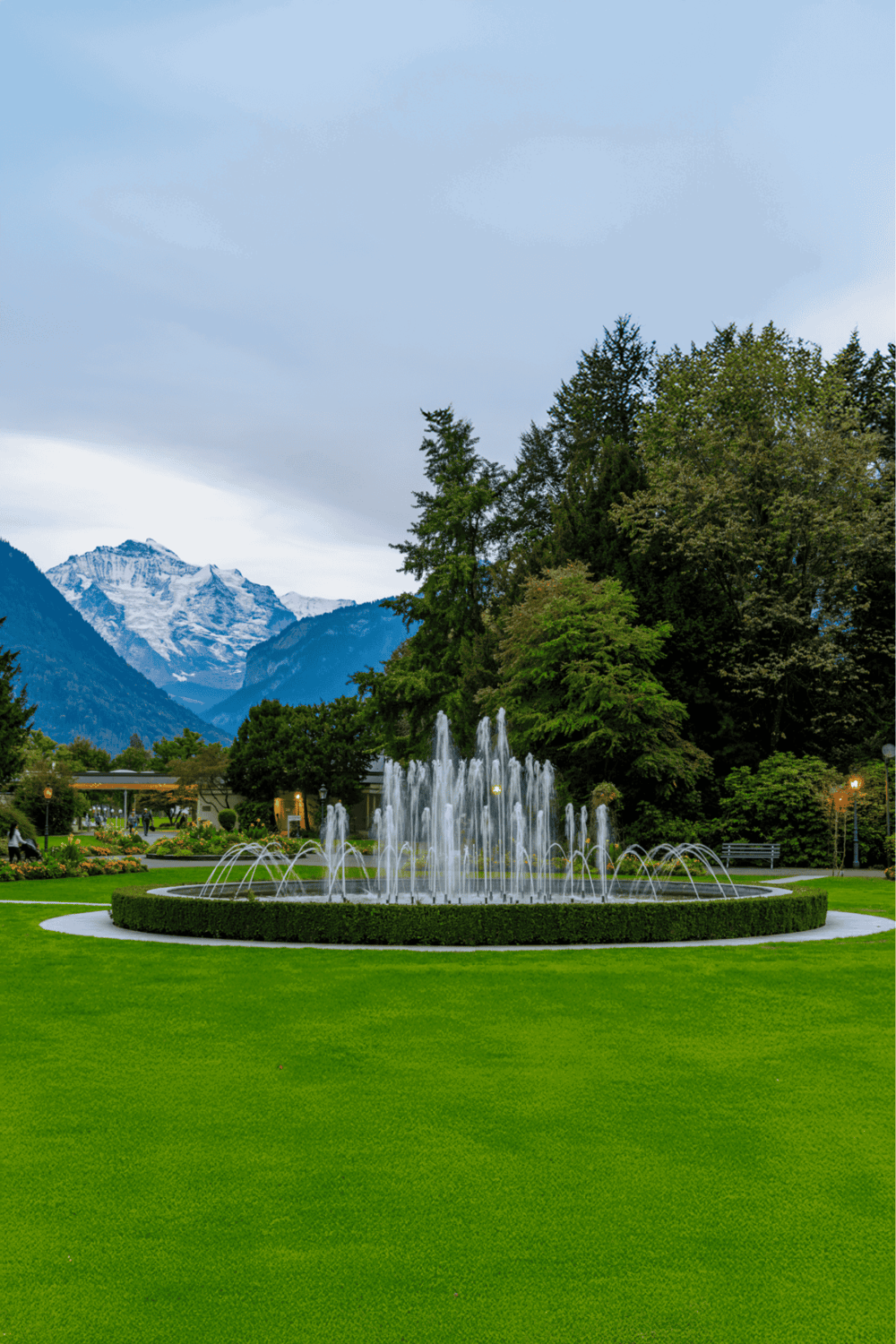 Vibrant park fountain surrounded by lush greenery with snow-capped mountains in the background.