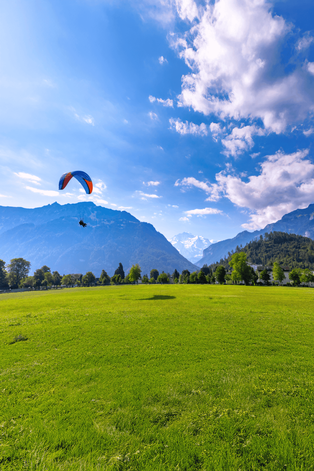 Colorful paragliding over green field with mountains and blue sky.
