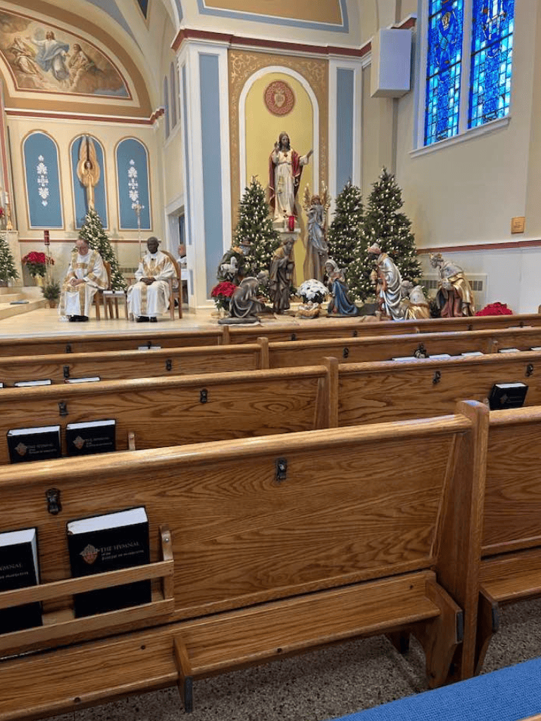 Nativity scene display inside a church during Christmas season, with priests, decorated altar, and stained glass windows.