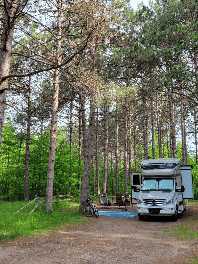 Secluded forest camping site with RV, bicycles, and outdoor seating in lush green woods.