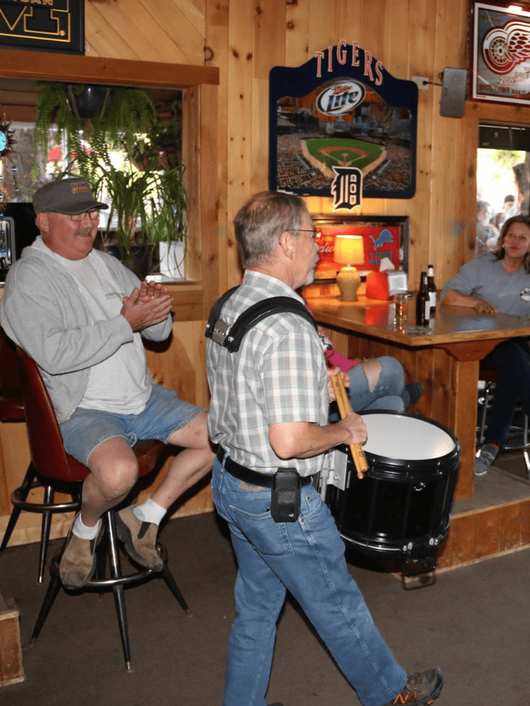 A man playing a drum while others watch in a cozy wooden bar setting with sports memorabilia and decor.