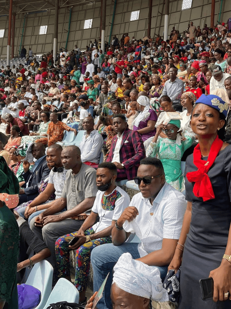 Vibrant crowd at a public event, diverse attendees in colorful traditional clothing and modern fashion, seated in stadium seating.