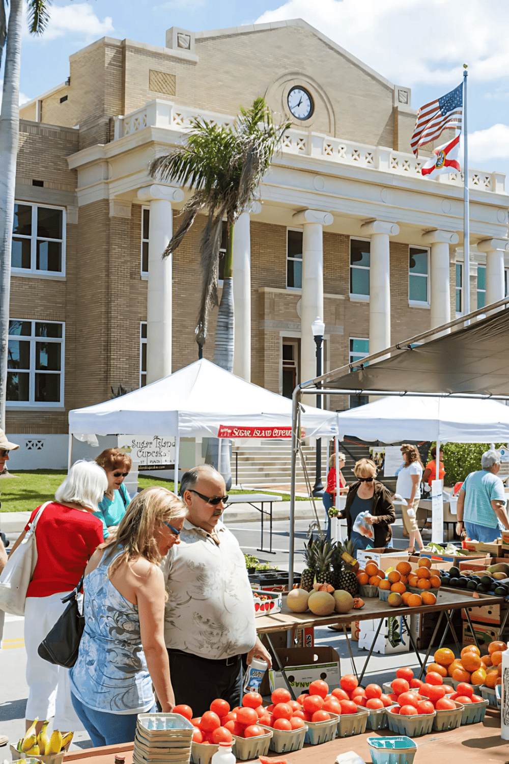 Bright daytime scene at a local farmers market in front of a historic building with a clock tower. Customers browse fresh produce and enjoy community atmosphere.