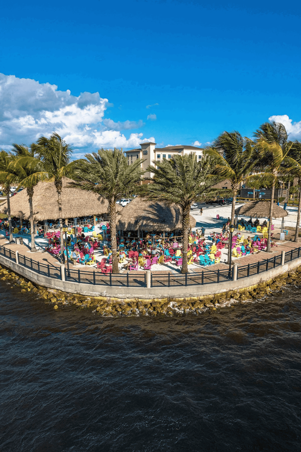 Relaxing waterfront tiki bar with colorful Adirondack chairs and palm trees, perfect for tropical vacation vibes.