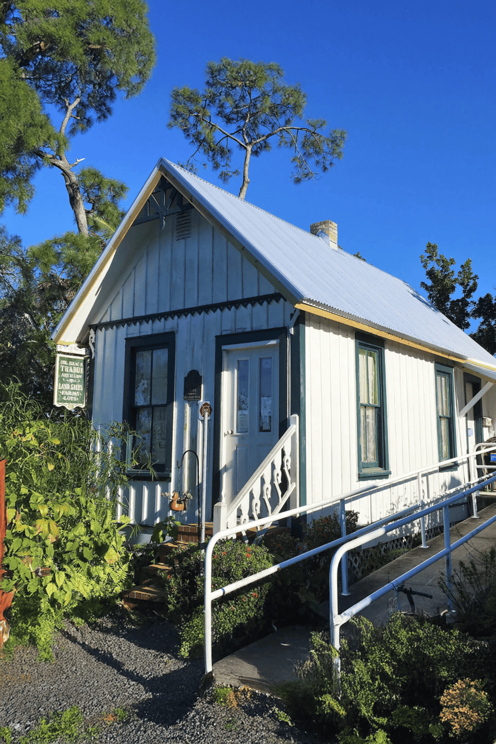 Coastal cottage with white siding and blue trim, situated among lush greenery under clear blue sky.