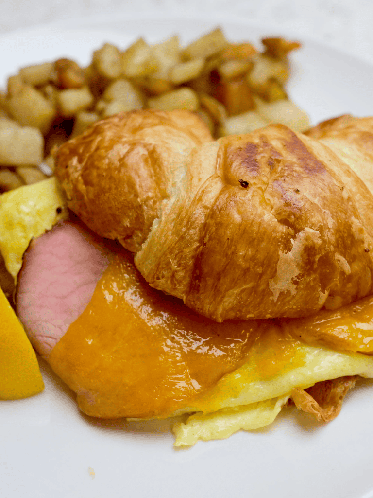 Scrambled eggs, croissant breakfast sandwich with cheese, ham, and hash browns on side.