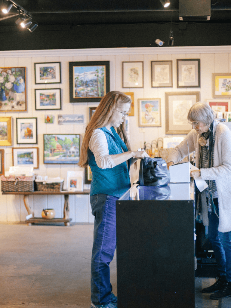 Art gallery shop with customers browsing and purchasing artwork, vibrant paintings on display, welcoming atmosphere, artistic shopping experience.