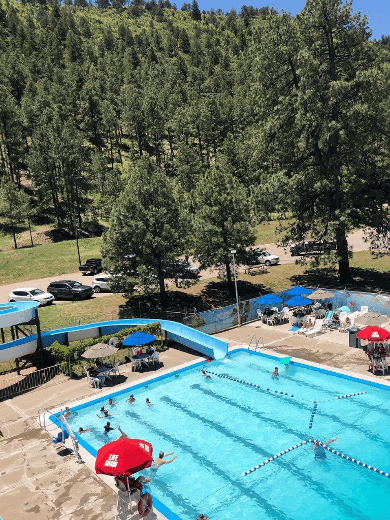 Sunny outdoor swimming pool with water slide and nearby shaded seating areas in a scenic forest setting.