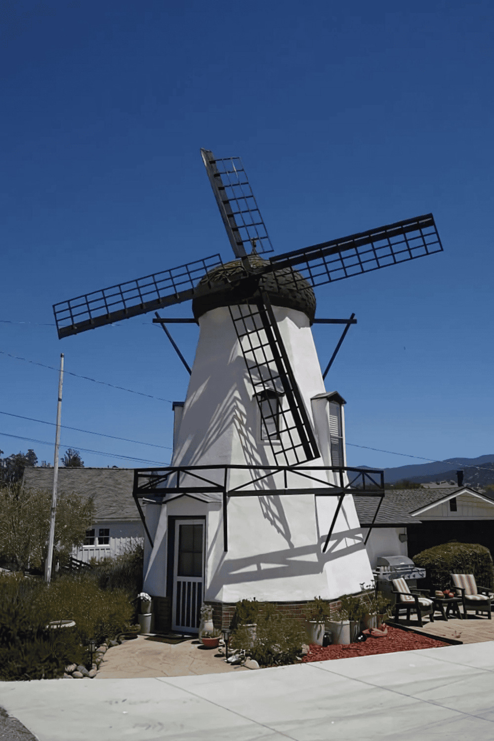 Windmill house with traditional blades and outdoor seating, located in a scenic area with mountains in the background.
