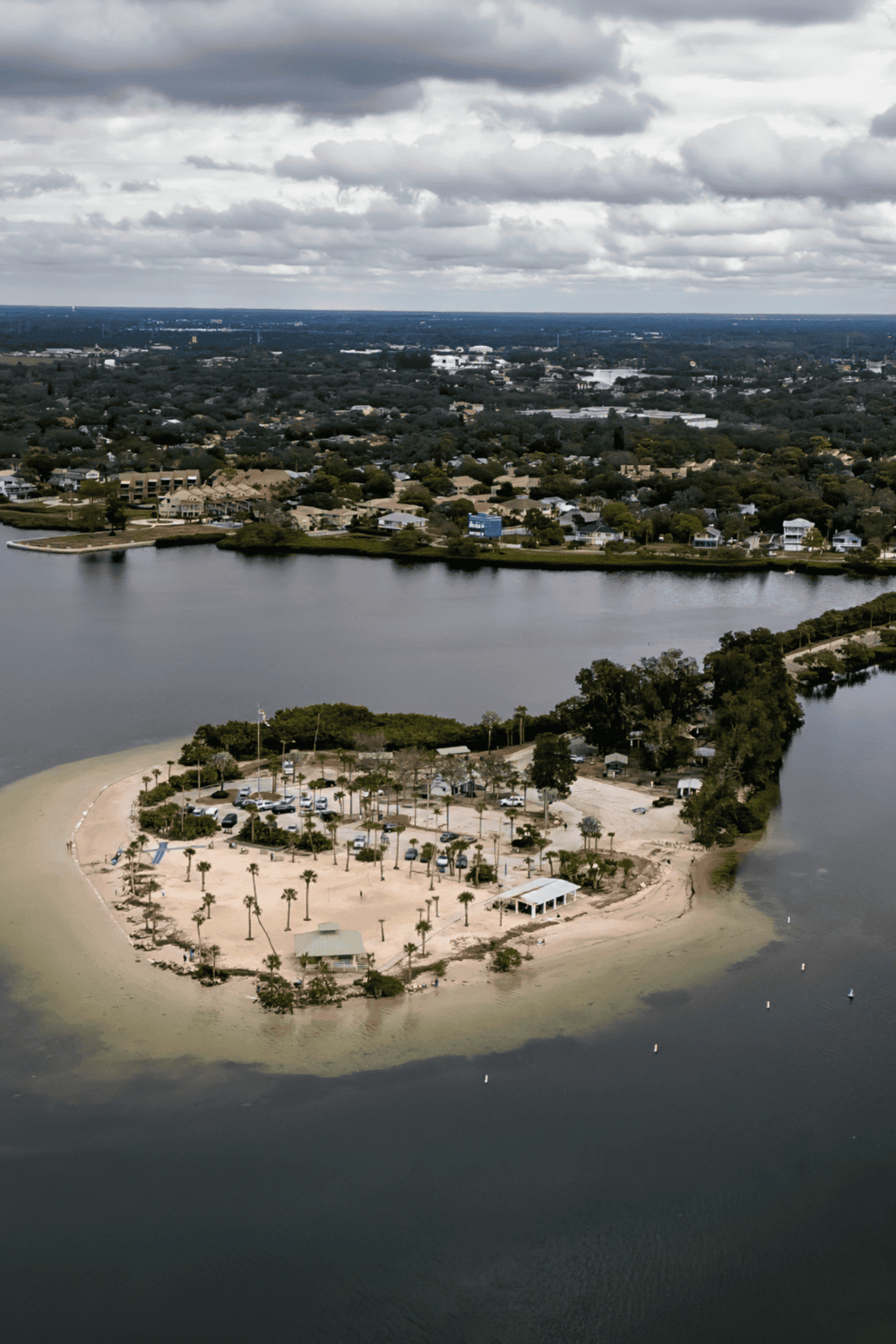 Seaside park on a small island with palm trees, water, and a parking lot under cloudy skies.