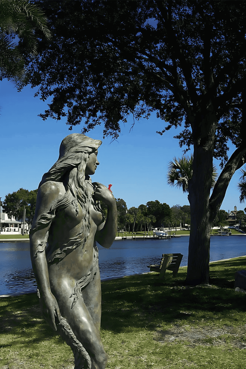 1. Serene waterfront park with statue of mermaid, lush trees, and boats in the background.
