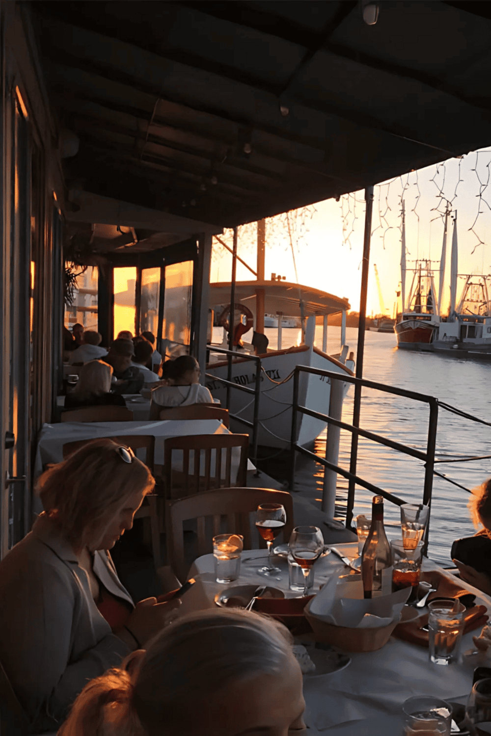 Outside dining at sunset on a boat cruise, with people enjoying drinks and views of yachts and water.