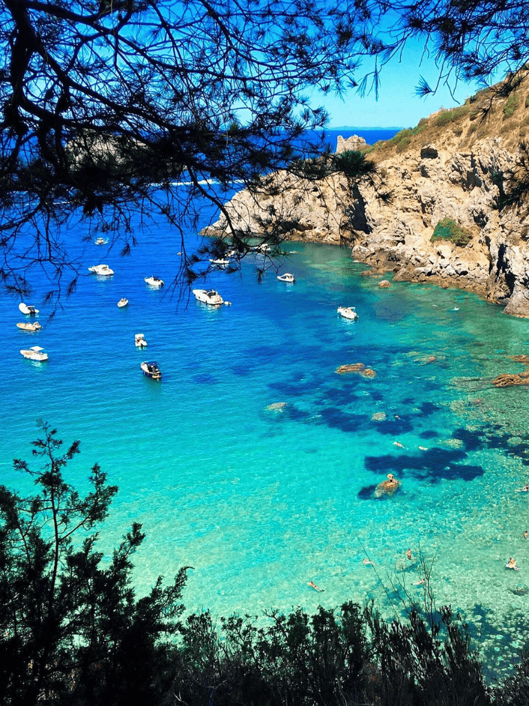 Blue ocean with anchored boats near rocky coastline, framed by trees.