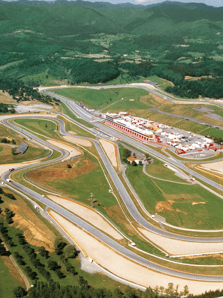 Wide aerial view of a complex race track circuit with multiple turns, surrounded by lush green mountains.