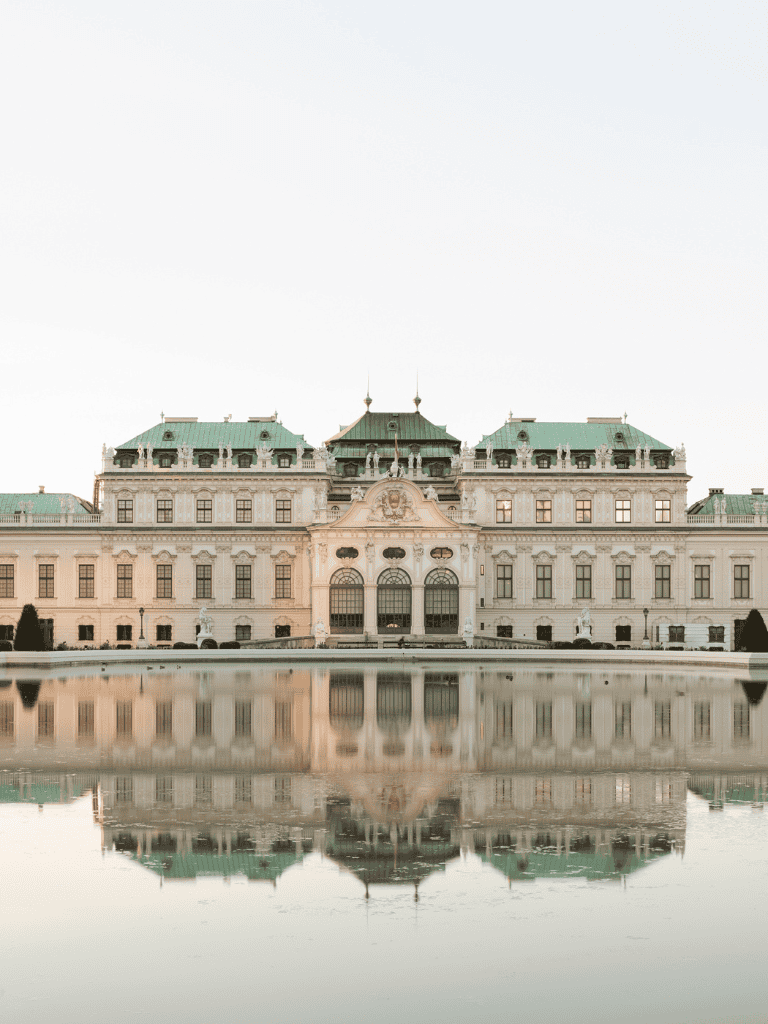 Luxurious European castle mirrored in water with ornate architecture and green rooftops.