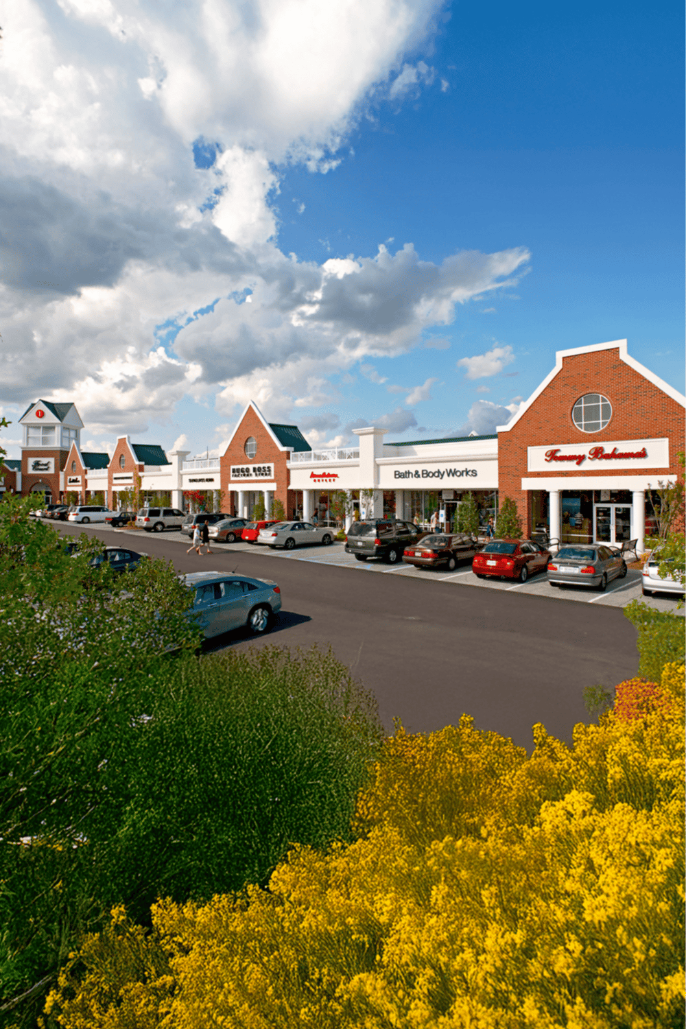 Bright shopping plaza with retail stores and parked cars under a partly cloudy sky.
