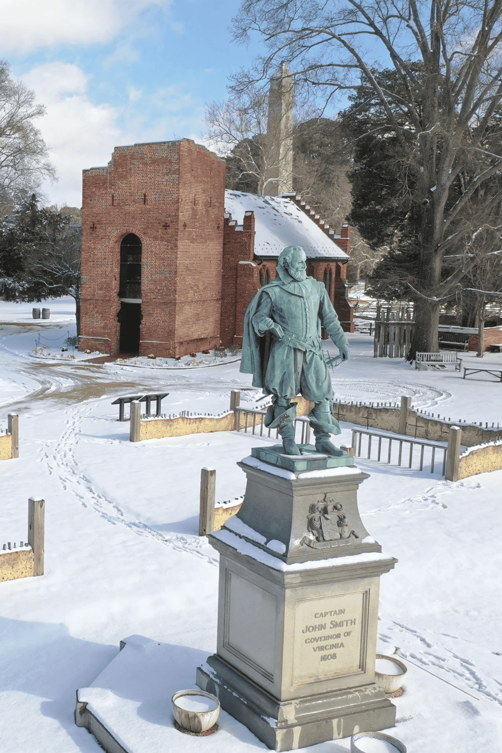 Statue of Captain John Smith in winter at Jamestown, Virginia, featuring historic architecture and snow-covered surroundings.