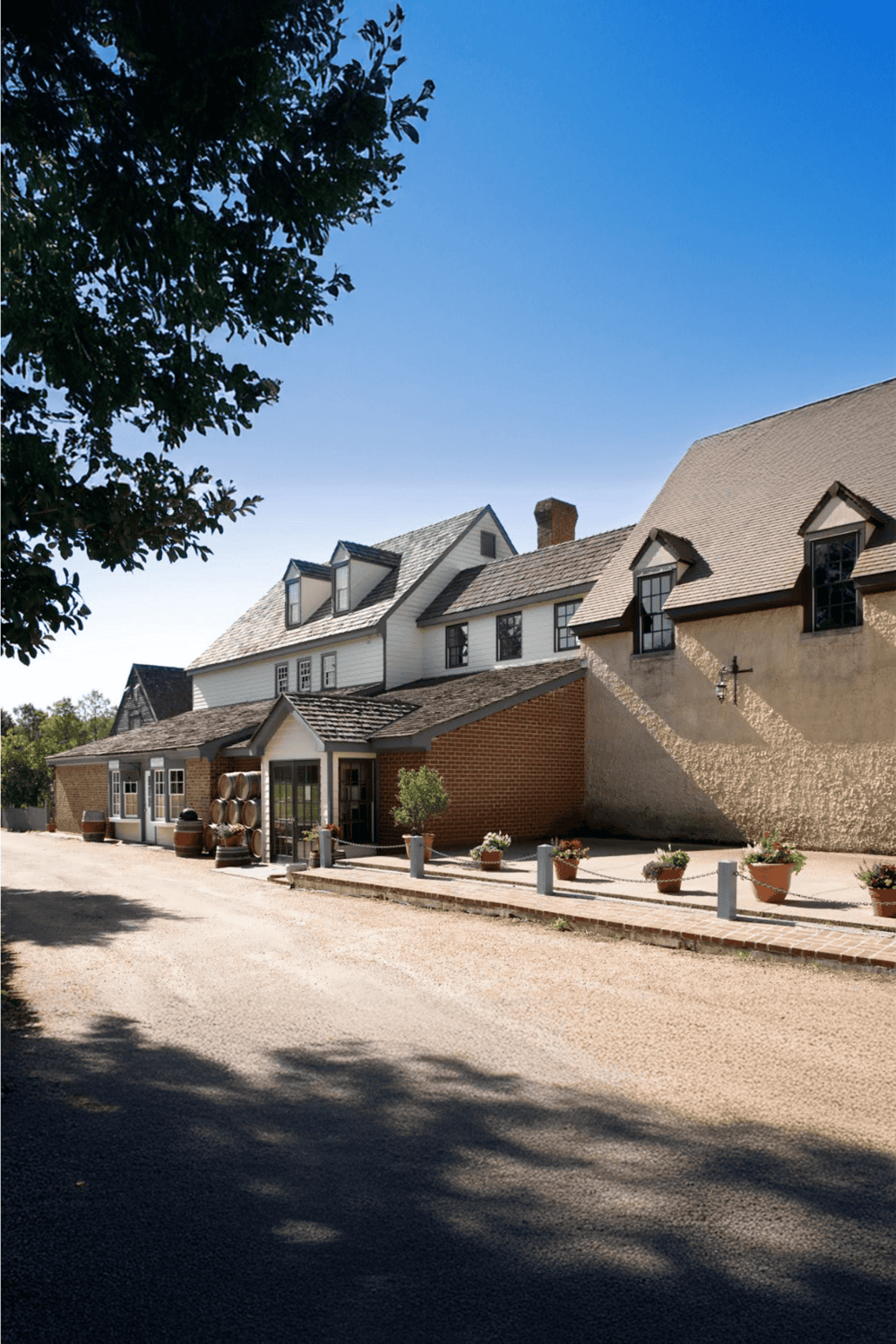 Cozy rustic cottage with brick and white siding, outdoor wine barrels, and potted plants under a clear blue sky.