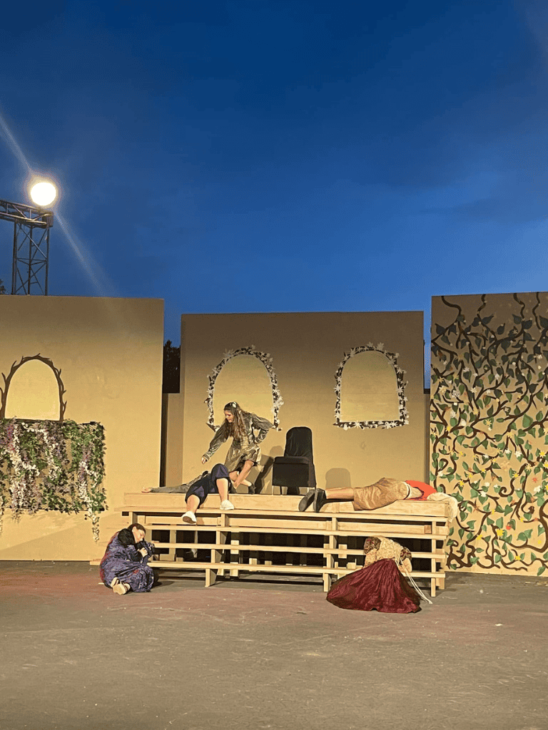 Elegantly staged theatrical performance on an outdoor stage with artistic backdrop and actors in costume.