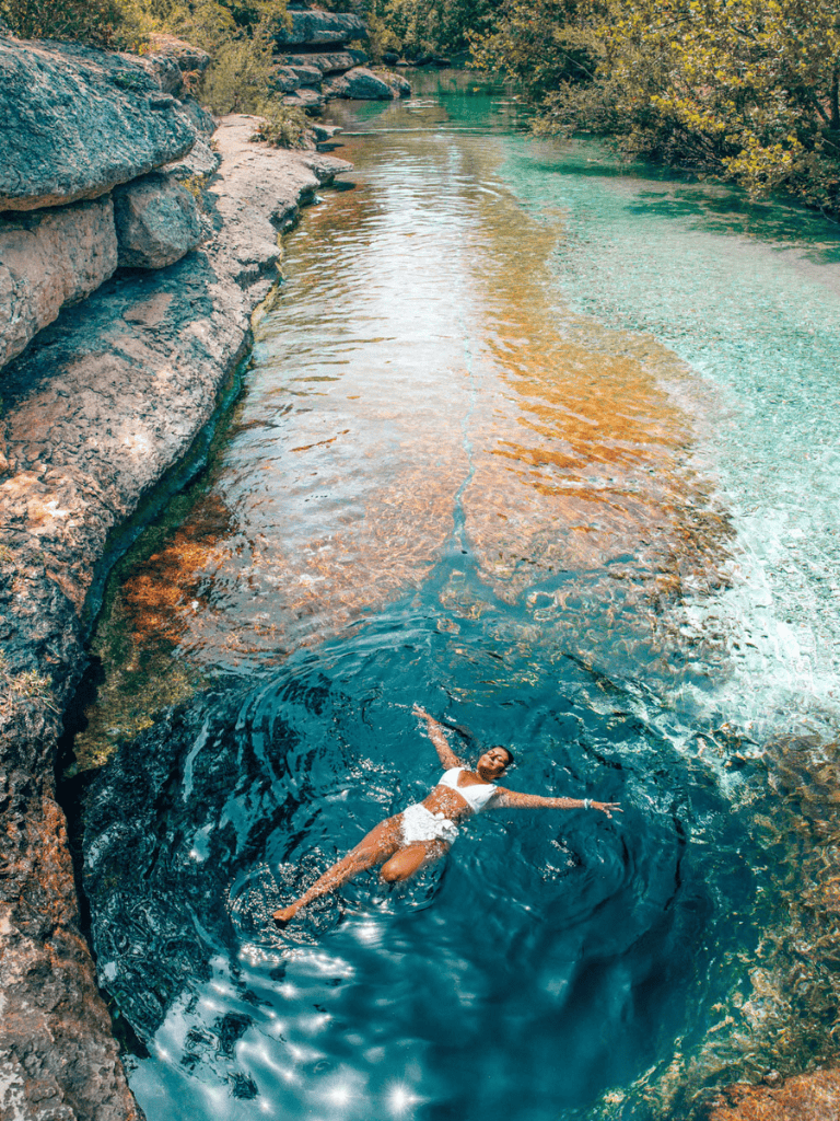 Peaceful woman floating in clear river surrounded by rocks and lush greenery.