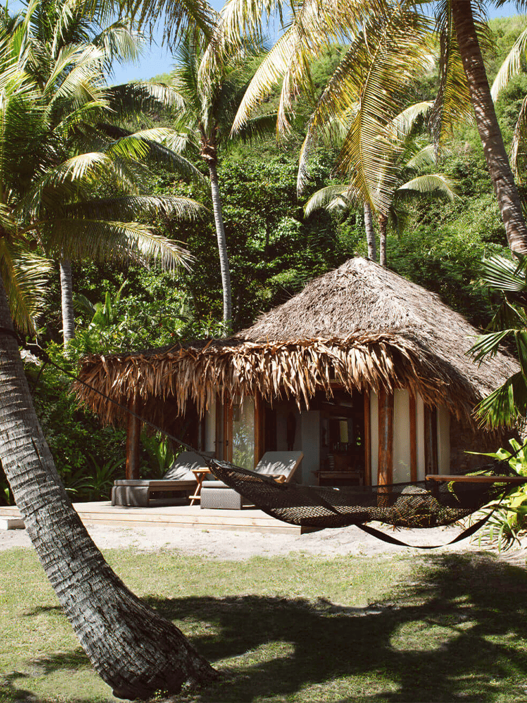 Tropical hut with thatched roof surrounded by lush palm trees and hammock, perfect for relaxing vacation.