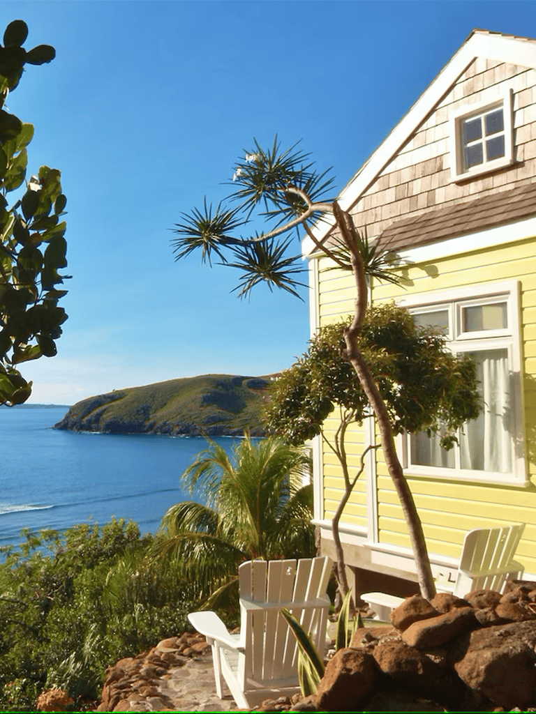 Aerial view of a cozy seaside house with ocean view, lush greenery, and outdoor seating in a tranquil coastal setting.