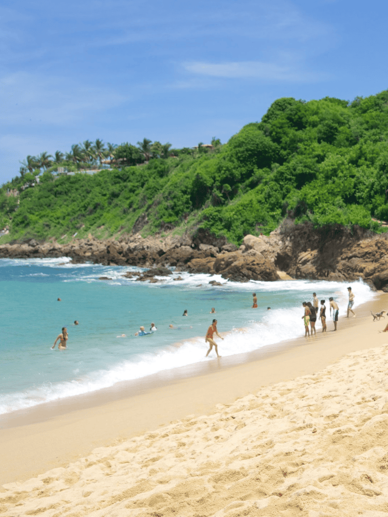 Beach with lush green hillside, people swimming and relaxing, scenic coastal view, tropical vacation destination.