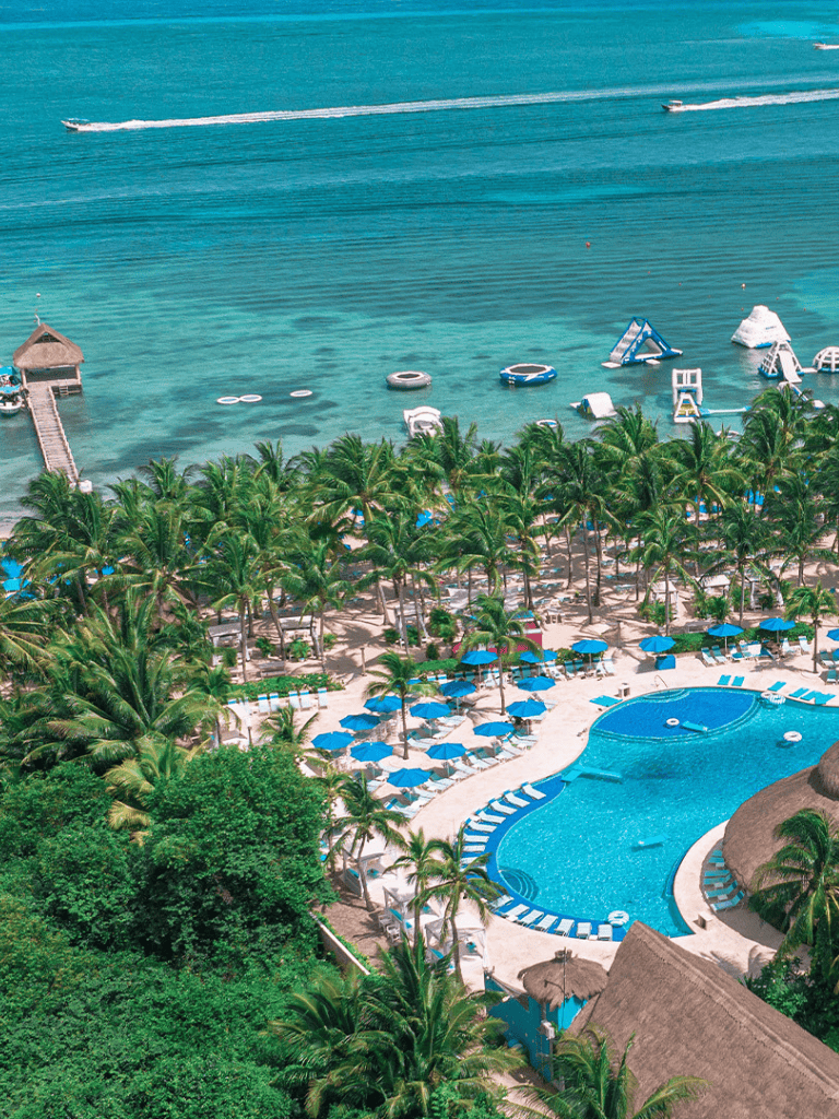Aerial view of tropical beach resort with pool, palm trees, and ocean water activities.