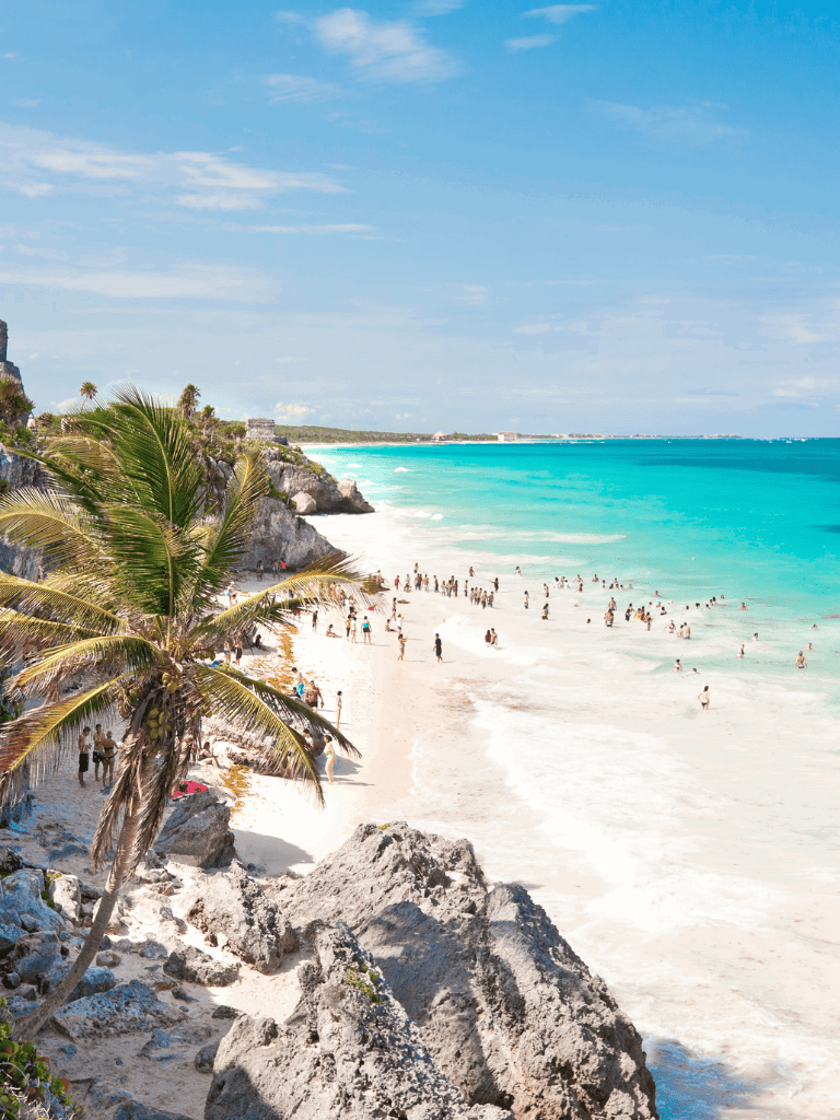 Tropical beach with turquoise waters, palm trees, and visitors relaxing and swimming in the ocean.