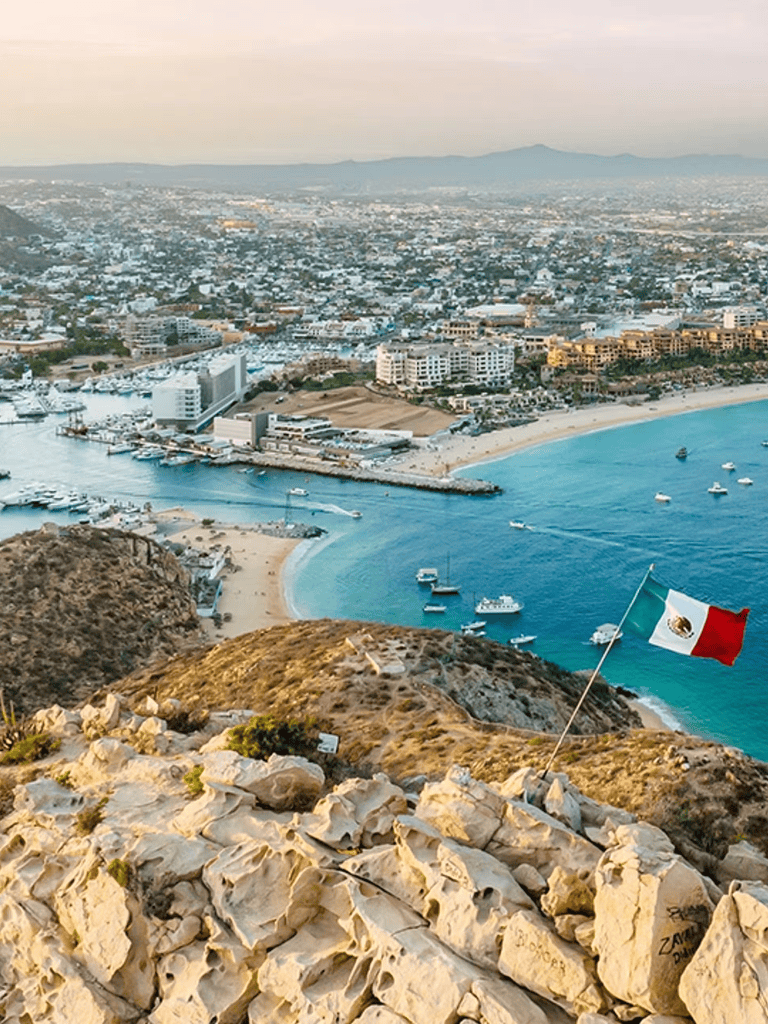 Panoramic view of Cancun coastline with marina and cityscape, featuring a Mexican flag on rocky foreground.