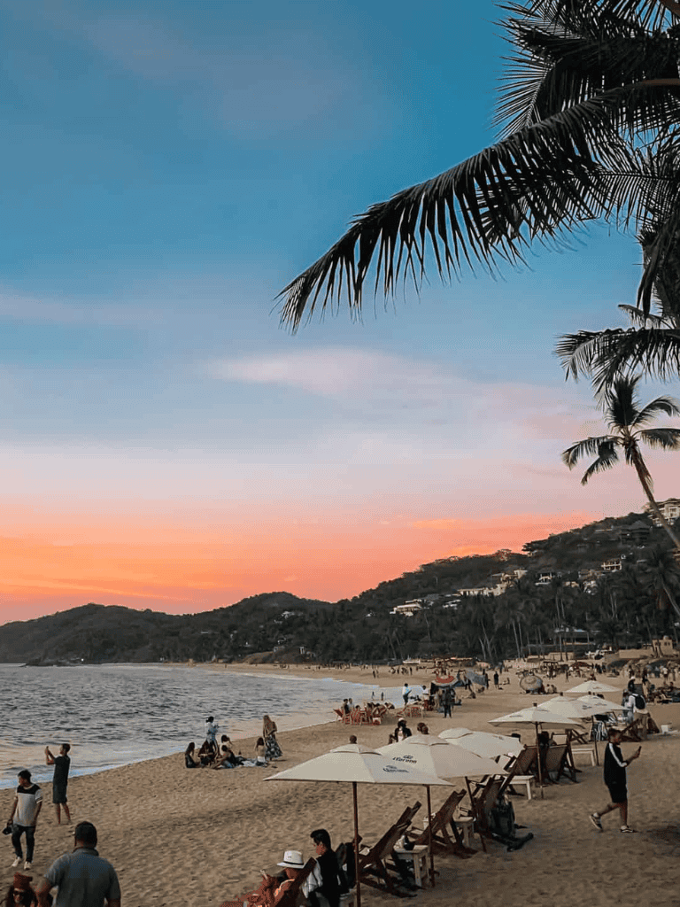 Sunset beach scene with palm trees, people relaxing under umbrellas, and scenic ocean view.