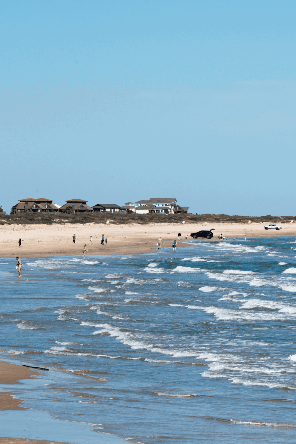 Bright sandy beach with ocean waves, houses in the background, and people enjoying coastal scenery, perfect for travel and vacation SEO.