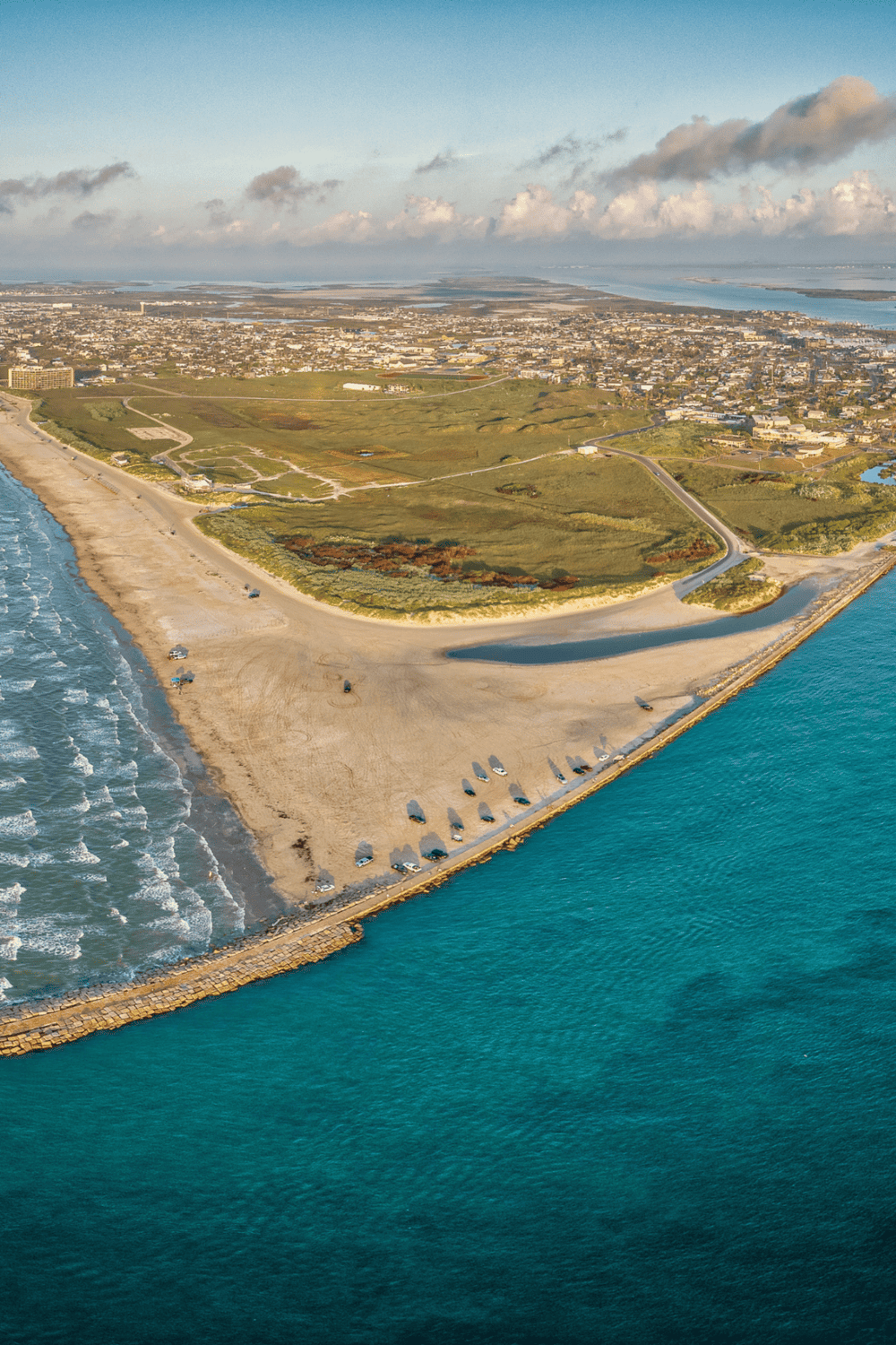 Aerial view of a coastal park with sandy beach, green landscapes, and city in the background, highlighting travel and outdoor destinations.