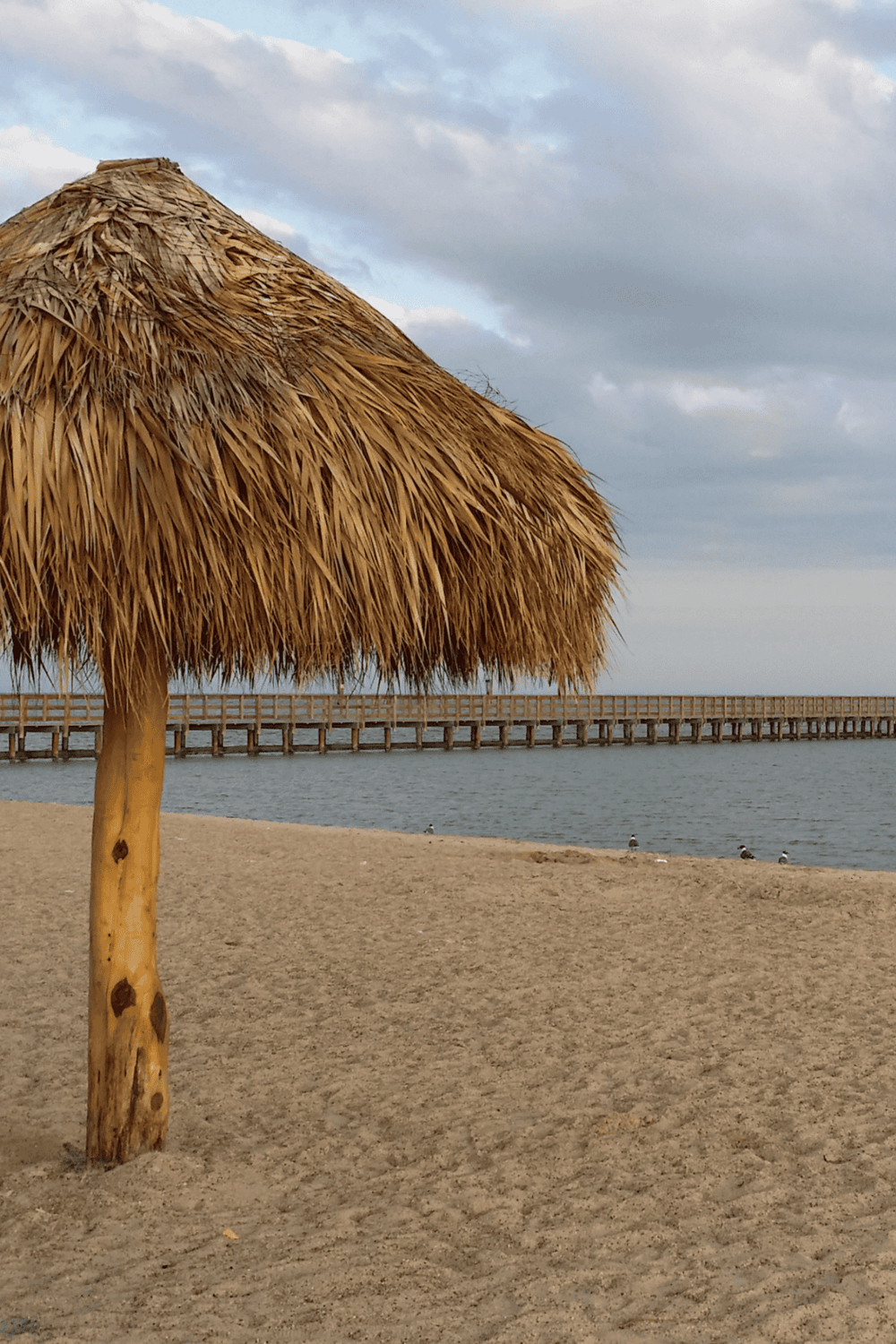 Relaxing beach scene with thatched umbrella, sandy shore, and pier at sunset.