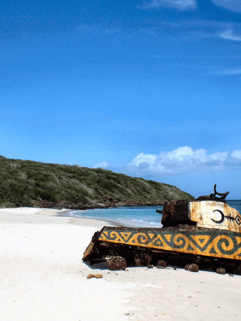 Rusty shipwreck on a tropical beach with vibrant ocean and lush green hills in the background.