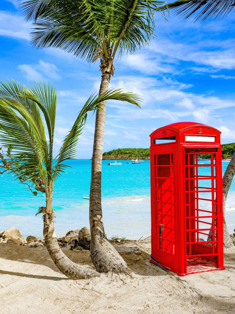 Bright red British-style telephone booth on tropical beach with palm trees and turquoise water.
