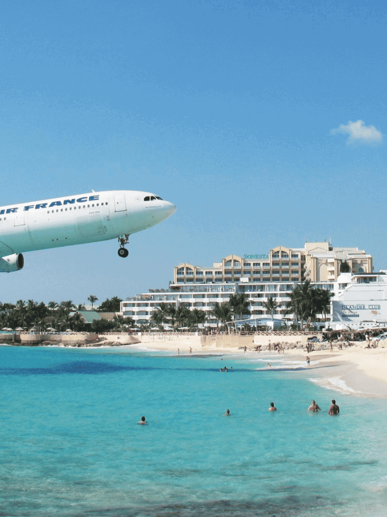 Airplane flying over Caribbean beach resort with clear turquoise water and hotel buildings in the background.