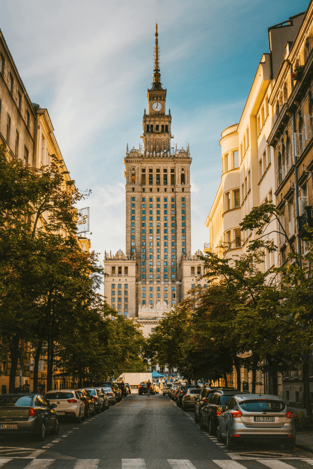 Tall historic building with clock tower at the end of a tree-lined street in a city.