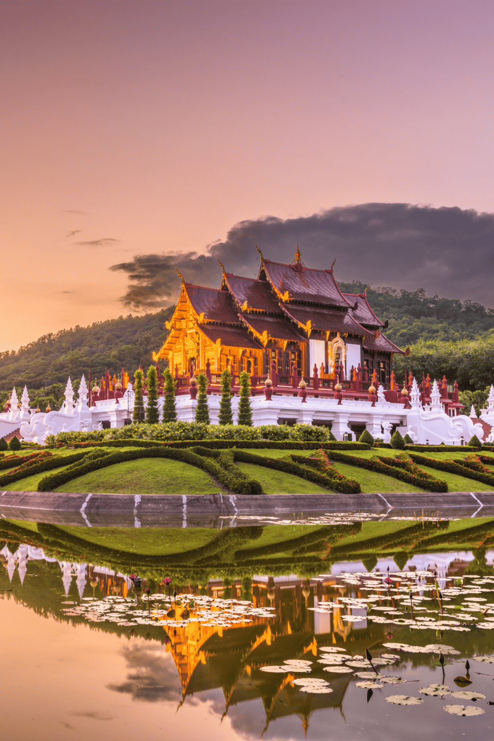 Traditional Thai temple at sunset with reflection in water, scenic religious site in Thailand, beautiful architecture, and lush mountain backdrop.