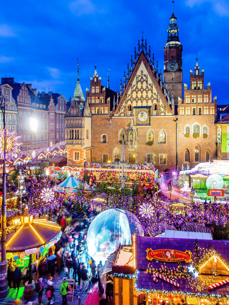 Colorful European-style Christmas market in historic city square at dusk, festive holiday decorations, and illuminated attractions.
