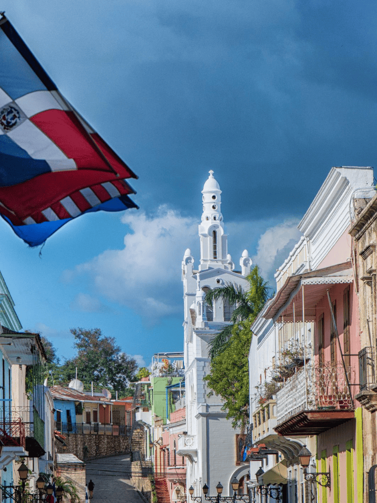 Colorful hillside street with historic buildings and a prominent white church tower under a partly cloudy sky.