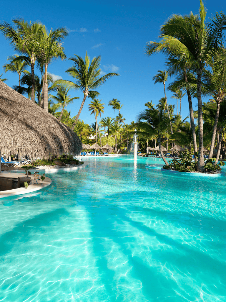 Luxurious tropical pool with palm trees and thatched roof cabanas beneath clear blue sky.