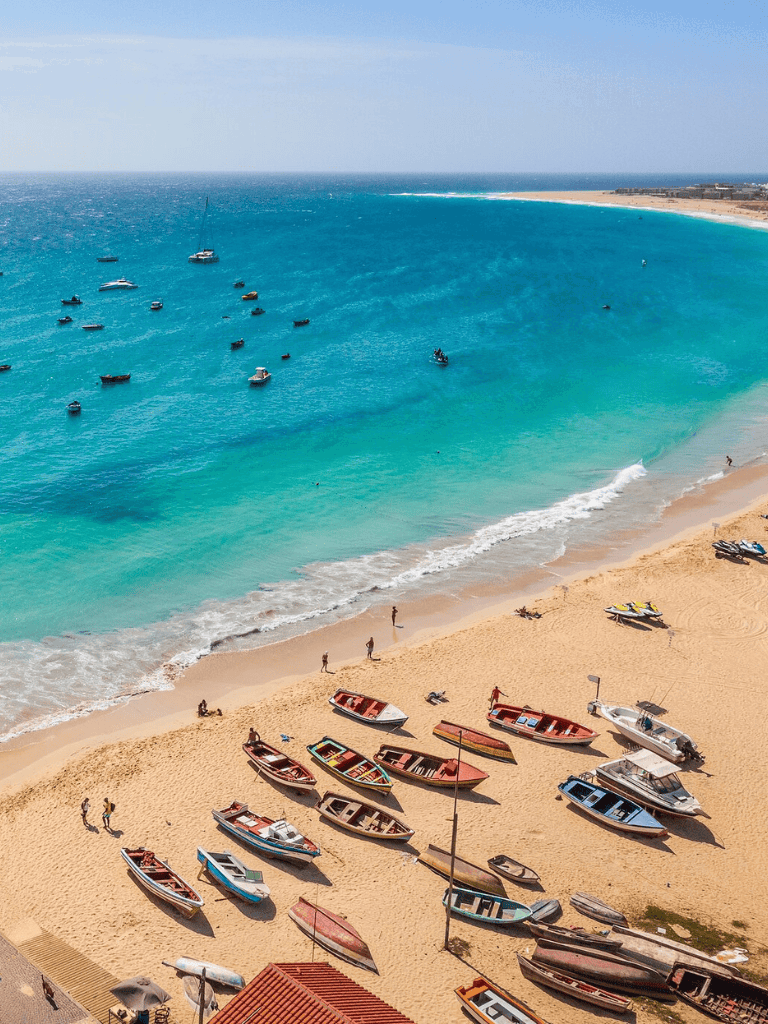 Boat harbor with colorful boats on sandy beach, turquoise water, lifeguards, and scenic ocean view.