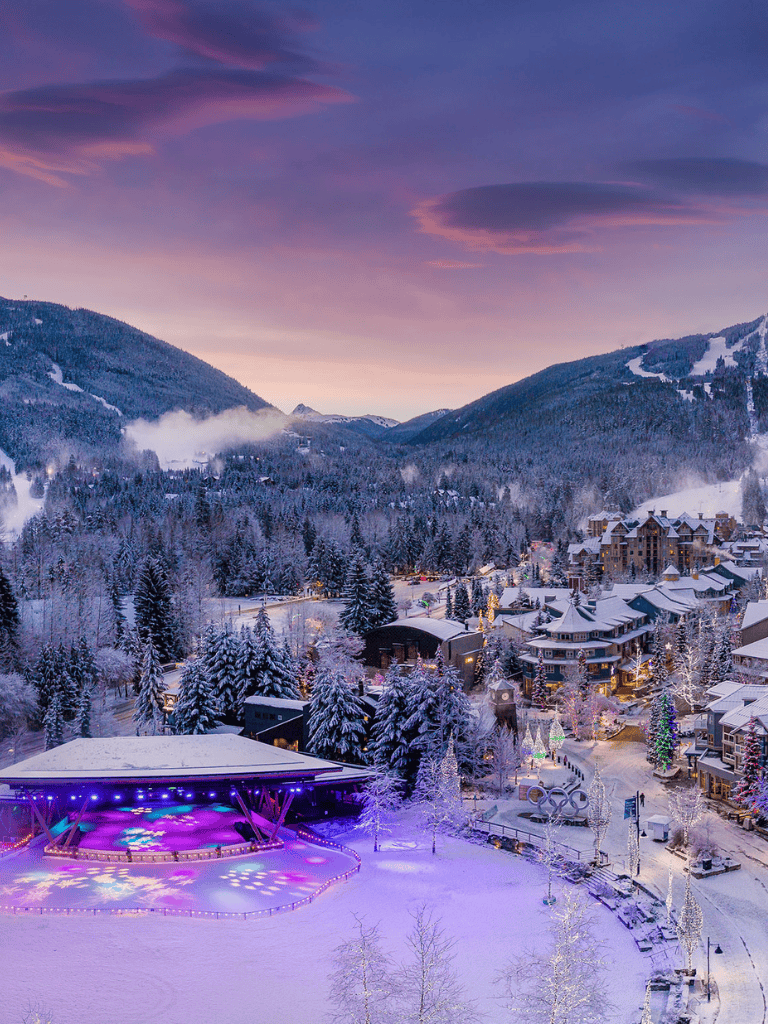 Snowy mountain village with an outdoor ice skating rink at dusk, winter lights, and ski slopes in the background.