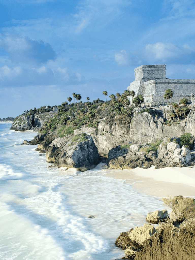 Ancient Mayan ruins on a coastal cliff with ocean waves, tropical trees, and blue sky in the background.