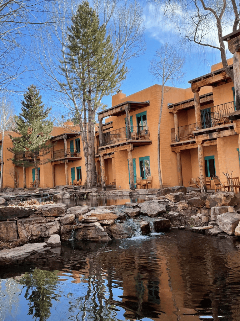 Charming adobe-style hotel with balconies and a scenic pond surrounded by rocks and trees.
