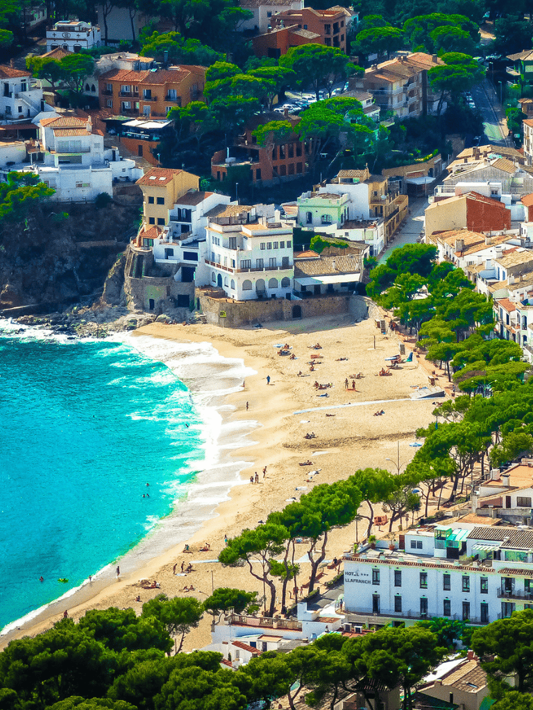 Colorful coastal hillside with beach and houses in Mediterranean style, lush green trees, clear turquoise water, scenic seaside view.
