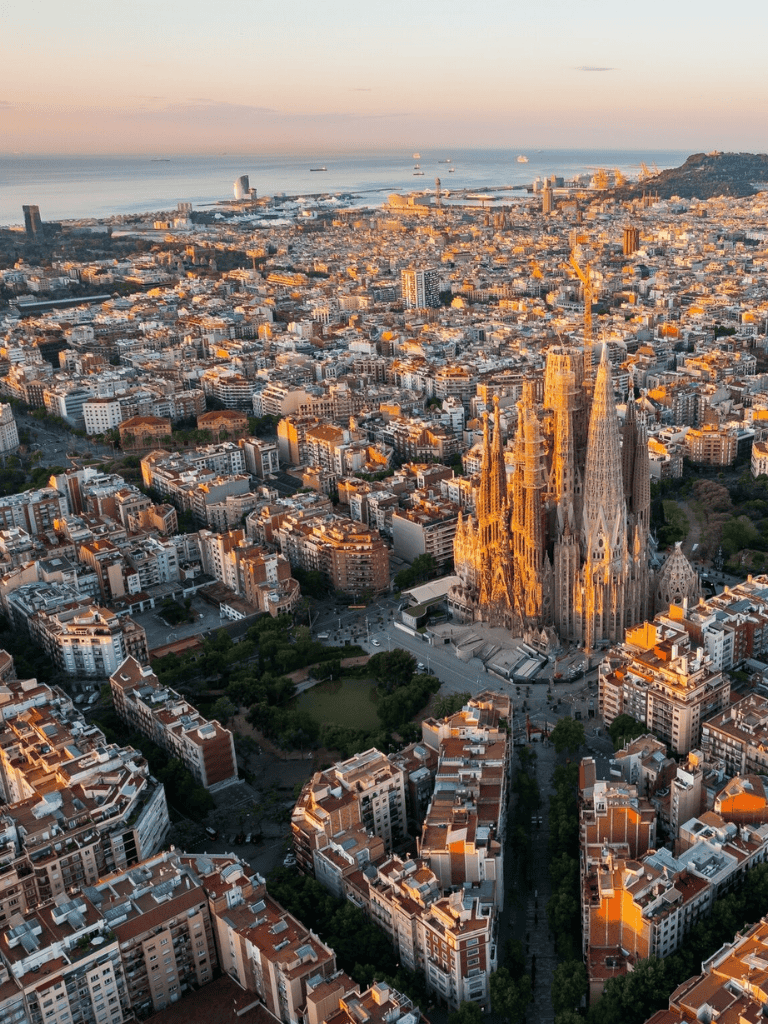 Tall city skyline with Sagrada Familia, Barcelona harbor, and coastal ships at sunset, showcasing Barcelona city's architecture and scenery.