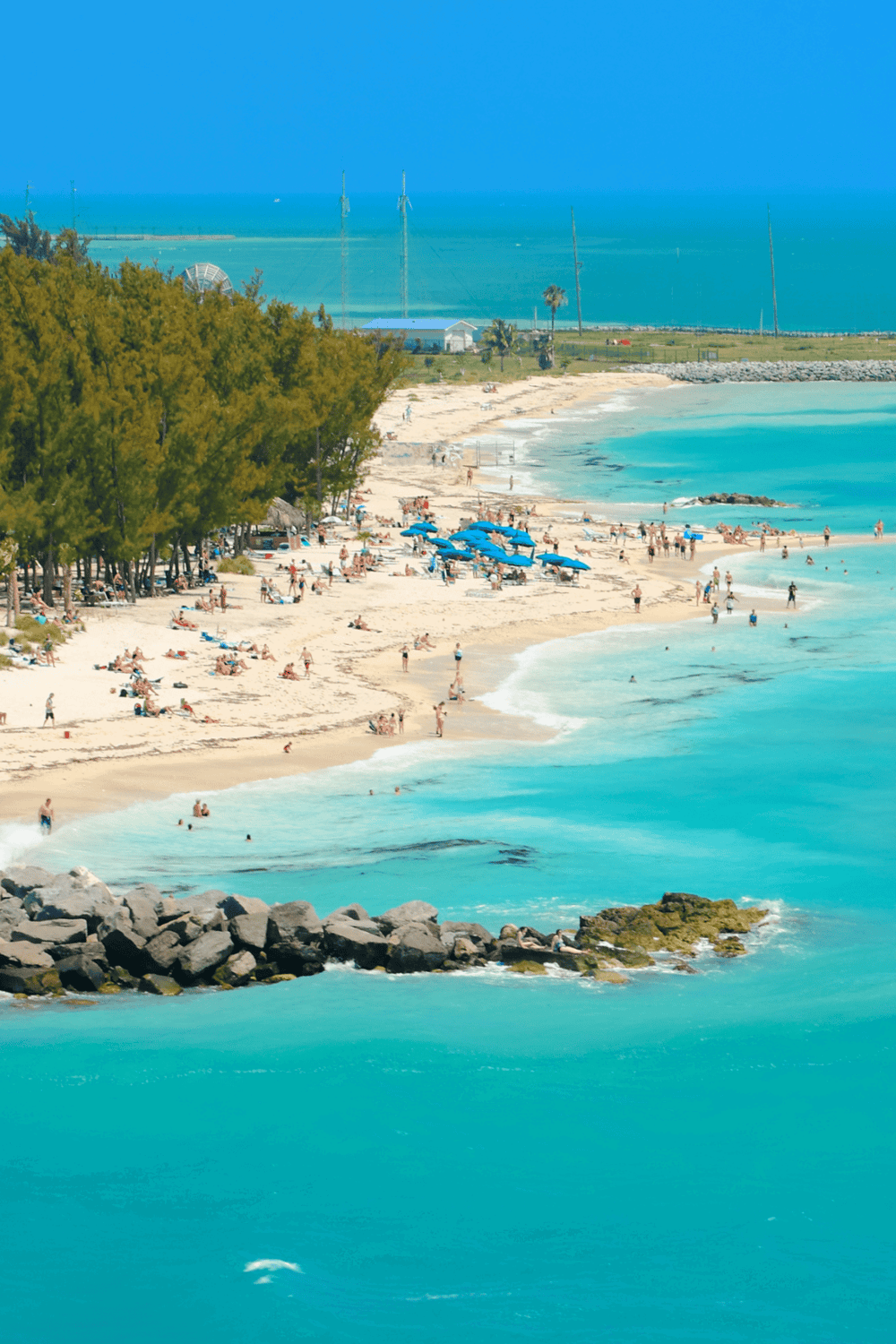 Vibrant beach with turquoise water, lush trees, and visitors sunbathing and swimming in clear ocean waters.