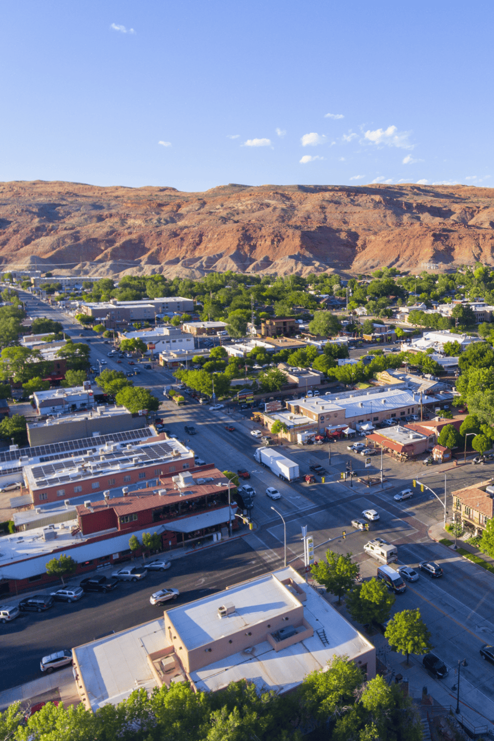 Aerial view of a cityscape with mountains in the background, sunny weather, and lush green trees along streets, showcasing urban environment and natural scenery.
